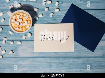 DANKESTEXT auf Danksagungskarte Inschrift positive Zitat Satz Grußkarte blauer Umschlag mit weißer Tasse Kaffee und Marshmallows auf holzblauem Hintergrund Urlaub. Draufsicht, flach liegend, minimalistisch Stockfoto