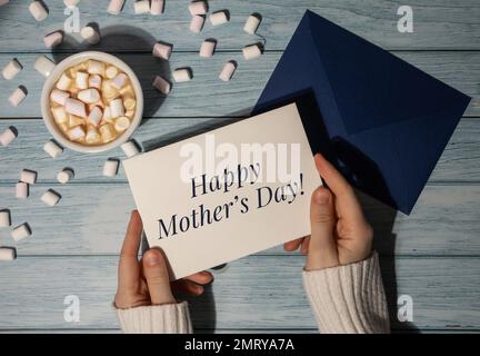 HAPPY MOTHER's DAY Text auf Karte Beschriftung positive Zitat Satz weibliche Hände halten Grußkarte blauer Umschlag mit weißer Tasse Kaffee und Marshmallows auf blauem Holz Hintergrund. Ansicht von oben, flach liegend, minimalistisches Branding-Konzept Stockfoto