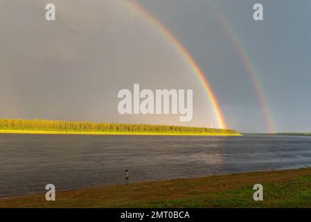 Ein Fischermädchen steht am Ufer eines Flusses mit einem großen doppelten Regenbogen vor dem Hintergrund eines Waldes und eines blauen Himmels. Stockfoto