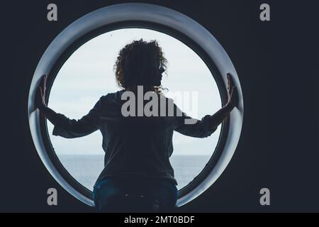 Porträt im dunklen Schatten eines Passagiers, der auf einem Bullaugenfenster sitzt und die Fahrt genießt. Reisemenschen denken an den Lebensstil. Eine Frau auf Kreuzfahrt mit Stockfoto