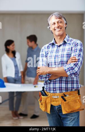 Lächelnder, erfahrener Architekt mit Kunden im Hintergrund. Porträt eines erwachsenen Architekten, der mit gefalteten Händen lächelt, während Kunden im Hintergrund diskutieren. Stockfoto