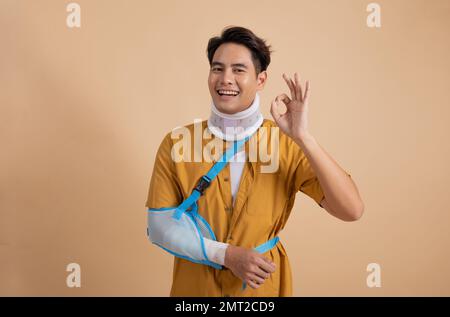 Attraktiver junger asiatischer Mann mit gebrochenem Arm in weicher Schiene, schmerzender Arm mit OK-Zeichen auf beigem Hintergrund isoliert, Unfallversicherungskonzept. Stockfoto