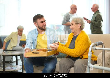 Junger Mann, der Seniorenessen im Geriatehospiz serviert. Seniorenfürsorge Stockfoto