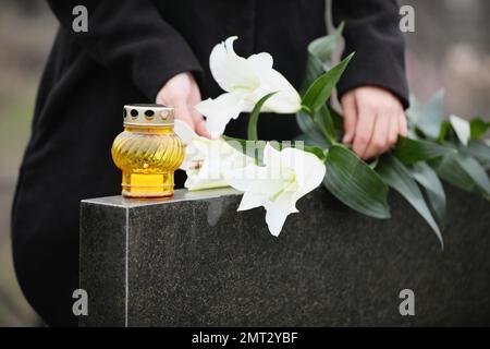 Eine Frau, die weiße Lilien in der Nähe eines Grabsteins aus schwarzem Granit hält, mit Kerzen im Freien, Nahaufnahme. Beerdigungszeremonie Stockfoto