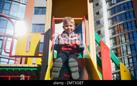 Ein kleiner Junge, der von der Rutsche runtergeht, lächelt, zeigt Daumen nach oben auf dem Spielplatz. Kleiner Winkel von niedlichem Mann oben auf der Rutsche. Moderne Wohngebäude im Hintergrund. Stockfoto