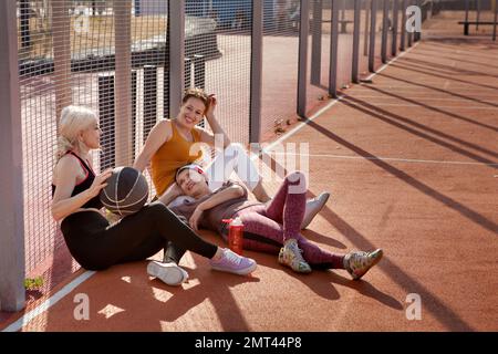Glückliche, reife Frauen entspannen sich auf dem Platz nach dem Basketballspiel. Weibliche Freunde reden, lachen und spielen draußen Sport Stockfoto