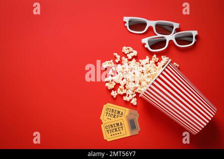 Umgekehrter Pappbecher mit köstlichem Popcorn, Kinokarten und 3D-Brillen auf rotem Hintergrund, flach liegend. Platz für Text Stockfoto