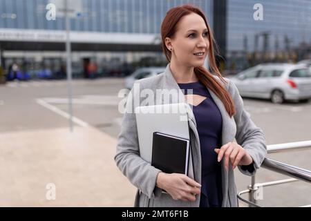 Reife rothaarige Geschäftsfrau mit einem Laptop in der Hand auf dem Parkplatz eines Business Centers Stockfoto