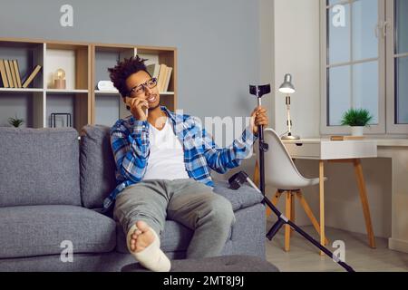Attraktiver junger Mann mit gebrochenem Bein, der auf dem Sofa sitzt und am Handy telefoniert Stockfoto