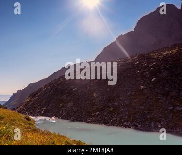 Die helle Sonne scheint mit Strahlen hinter einem Steinberg neben dem türkisfarbenen Wasser des Flusses Karakabak. Stockfoto