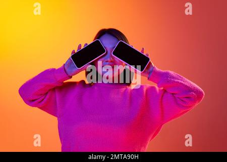 Technische Geräte, moderne Technologien. Ein junges Mädchen in pinkfarbenem Pullover, das mit zwei Mobiltelefonen posiert, auf einem abgestuften orangefarbenen Hintergrund in Neonlicht. Konzept von Stockfoto