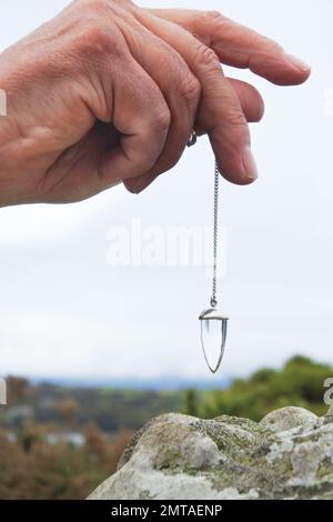 Männliche Hand mit einem Kristallpendel, die über einen stehenden Stein taucht - John Gollop Stockfoto