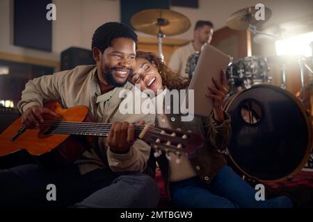 Musiker, Gitarre und ein Tablet für Musik, E-Mails und Klangerfahrung in einem dunklen Studio. Fröhlich, lesend und schwarzer Mann und Frau, die weiterströmen Stockfoto