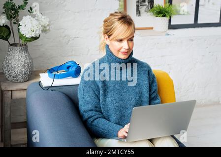 Attraktive blonde Frau, die zu Hause sitzt und ein Notebook für die Arbeit benutzt. Lächelnde Frau mittleren Alters in lässiger Kleidung. Stockfoto