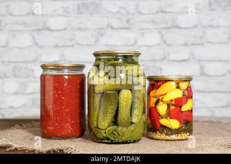 Hausgemachte Konserven. Eingelegte Snacks für den Winter. Eingelegte Gurken, italienische Tomaten im eigenen Saft und Mini-Chili-Paprika in Glasgefäßen. Stockfoto