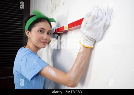 Asiatische junge Hausfrau im Handschuh mit einer Wasserwaage an der Wand Stockfoto
