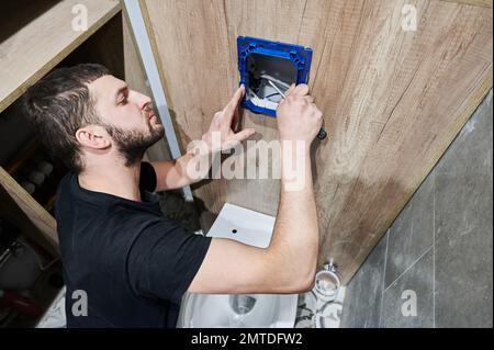 Bärtiger Mann, der ein Schraubendreher hält, während er die Toilettenspülung in einer Wohnung installiert, die gerade renoviert wird. An der Wand montierter Klempner-Einstellrahmen für Toilettenbetätigungselemente. Stockfoto