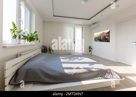 Graue Decke auf dem Bett mit hölzernen bedhead in einfachen Schlafzimmer Einrichtung mit dunklen Poster und Stuhl unter dem Fenster Stockfoto