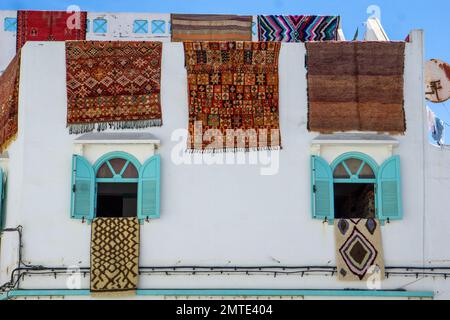 Weiße Fassade in Maroc mit hellblauen Fenstern, die weit geöffnet sind. Bunte Teppiche hängen an einem sonnigen Tag mit blauem Himmel vom Dach und den Fenstern. Stockfoto