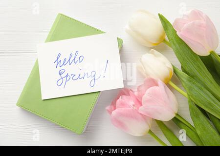 Karte mit den Worten „HELLO SPRING“, Notizbuch und Tulpen auf weißem Holztisch, flach liegend Stockfoto