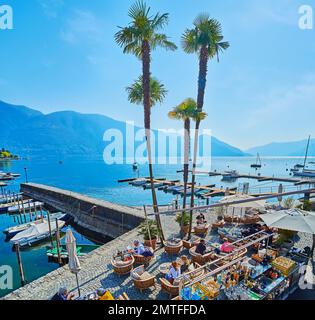 Das Seeufer von Ascona mit gemütlichen Restaurants am Lago Maggiore mit Blick auf Schiffswerften mit festgefahrenen Fischerbooten und Alpenlandschaft im Hintergrund, wie Stockfoto