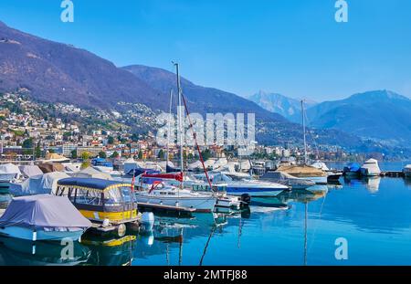 Die Linie von festgemachten Booten und Yachten auf dem Lago Maggiore mit alpinen Hängen und kleinen Häusern im Hintergrund, Locarno, Schweiz Stockfoto
