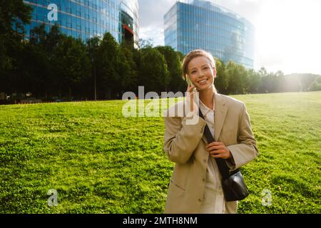 Junge, schöne lächelnde Geschäftsfrau, die telefoniert und zur Seite schaut, während sie auf dem sonnigen Rasen in der Stadt steht Stockfoto