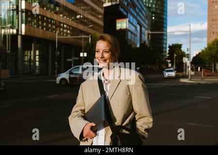 Junge, schöne, glückliche Geschäftsfrau mit einem Notebook, das zur Seite blickt, während sie auf der Straße spaziert Stockfoto