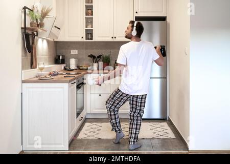 Ein bärtiger Mann in Pyjamahose und ein weißes T-Shirt tanzt morgens mit Kopfhörern in der Hausküche, während er Kaffee kocht. Stockfoto