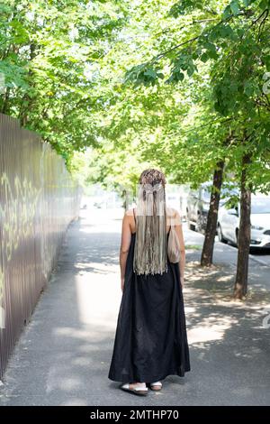 Ein Mädchen in schwarzem Kleid und langen Afro-Zöpfen läuft auf einer Sommerstraße, Rückansicht Stockfoto