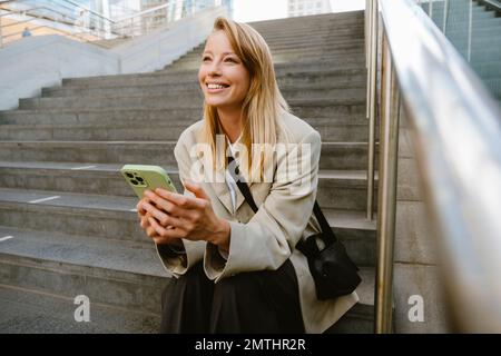 Junge weiße Frau, die lächelt und ihr Handy benutzt, während sie draußen auf der Treppe sitzt Stockfoto