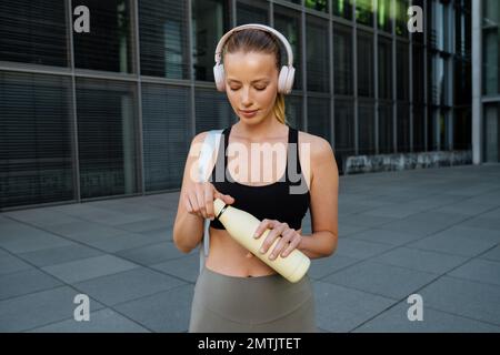 Weiße junge Frau mit Kopfhörern, die nach dem Training im Freien mit einer Wasserflasche herumläuft Stockfoto