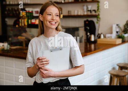 Blonde weiße Frau, die lächelt und ein Klemmbrett hält, während sie im Café drinnen arbeitet Stockfoto