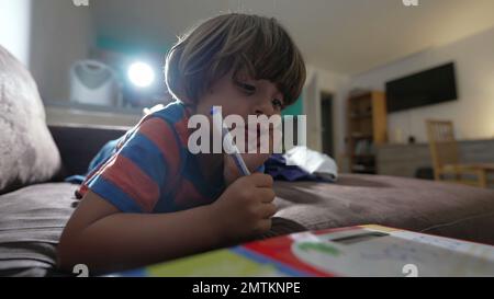 Ein aufmerksames Kind, das auf dem Sofa liegt und Hausaufgaben macht. Pensives Kind mit Hand-in-Kinn konzentrierter Gedankenlösung Stockfoto