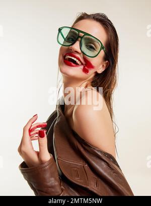 Roter Lippenstift ist umgereift. Hübsches junges braunes Mädchen mit nassem Haar in einer stilvollen Lederjacke, die über den grauen Studiohintergrund lächelt. Das Konzept der Schönheit Stockfoto