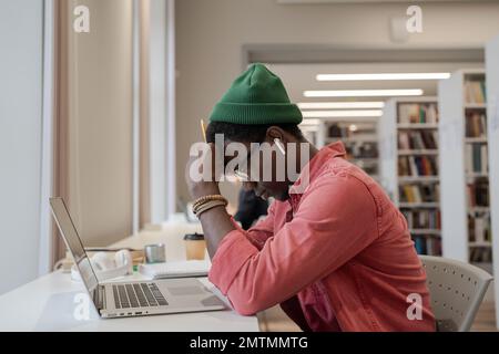 Ein müder, apathischer Afroamerikaner sitzt mit geschlossenem Laptop an einem Tisch in der Bibliothek Stockfoto