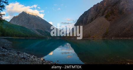 Panorama des Ufers des Lake Shavlinskoe mit Rauch eines Touristenfeuers im Schatten der Berge mit Reflexion von Gipfeln mit Gletschern Stockfoto