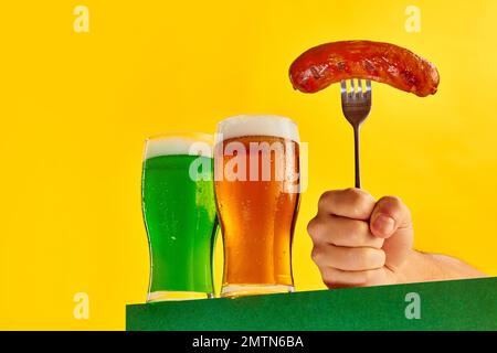 Zwei Gläser kühles Grün- und Lagerschaumbier und gegrillte Wurst auf einer Gabel auf gelbem Hintergrund. Konzept der St. patrick's Day Feier, Brauerei Stockfoto