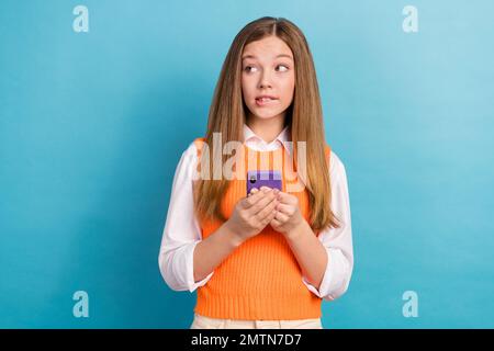 Foto von einem gedanklichen, nachdenklichen Mädchen, das Lippen beißt und das Telefon leer hält, isoliert auf blauem Hintergrund Stockfoto