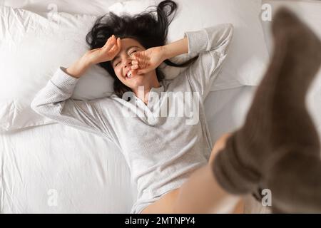 Glückliche junge europäische Dame schläft auf weißem Bett, hebt die Beine in Socken, wacht auf, genießt einen warmen, guten Morgen Stockfoto