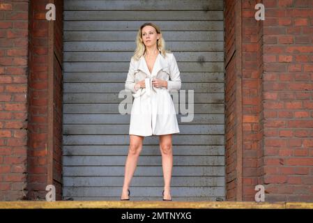 Eine attraktive blonde Frau mittleren Alters, die einen weißen Mantel oder Trenchcoat trägt und nichts darunter hat, steht vor einer grauen Rolltür eines alten B Stockfoto