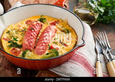 Omelette oder Frittata mit zwei Würstchen, geschmolzenem Käse, Kirschtomaten, Brokkoli und Petersilie auf roter Pfanne oder Eisenpfanne auf altem rustikalem Holzbacken Stockfoto