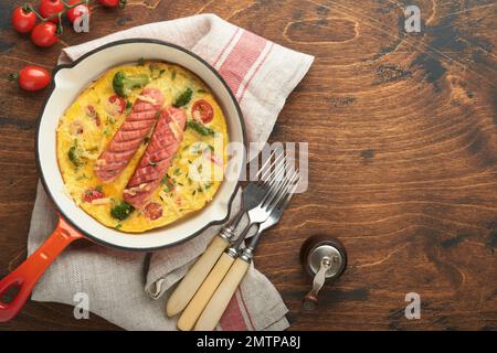 Omelette oder Frittata mit zwei Würstchen, geschmolzenem Käse, Kirschtomaten, Brokkoli und Petersilie auf roter Pfanne oder Eisenpfanne auf altem rustikalem Holzbacken Stockfoto