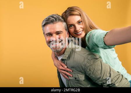 Wir haben Spaß zusammen. Glückliche Ehepartner mittleren Alters, die Selfie machen, Frau Huckepack, die auf dem Rücken ihres Mannes reitet Stockfoto