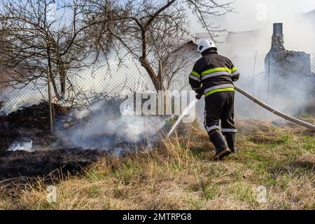 Frühlingsfeuer, brennendes trockenes Gras in der Nähe von Gebäuden auf dem Land. Der Feuerwehrmann löscht die Flamme. Umweltkatastrophe. Stockfoto