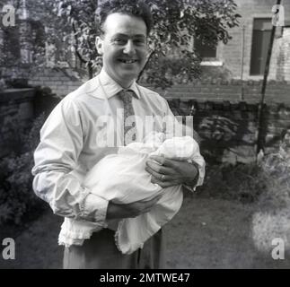 1950er, historisch, mit einem großen Lächeln im Gesicht, ein Vater hielt sein Baby in der Hand, gekleidet in einem Spitzenkleid, nach der Taufe, stand in einem Garten eines terrassenförmigen Hauses, Northumberland, England, Großbritannien. Stockfoto