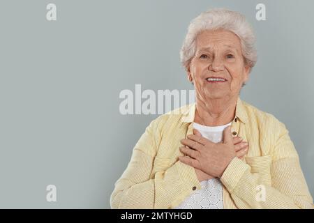 Dankbare ältere Frau mit Händen auf der Brust vor grauem Hintergrund. Platz für Text Stockfoto