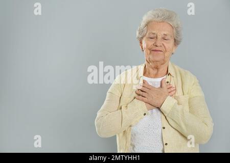 Dankbare ältere Frau mit Händen auf der Brust vor grauem Hintergrund. Platz für Text Stockfoto