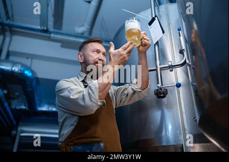 Ein Brauereiarbeiter, der sich frisch zubereitetes Bier in einem Glasbecher ansah Stockfoto