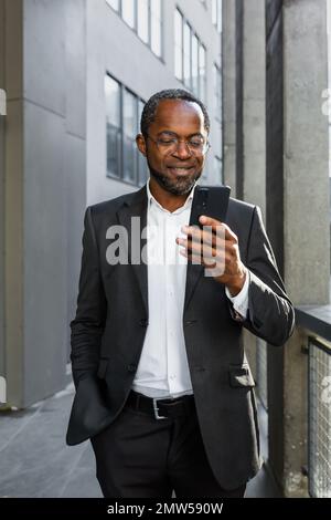 Vertikales Foto. Porträt eines lächelnden afroamerikanischen Geschäftsmanns in Anzug und Brille, der draußen neben einem Bürozentrum steht und seine Hand in der Tasche hält. Verwendet das Telefon, tätigt Anrufe, gibt Nachrichten ein. Stockfoto